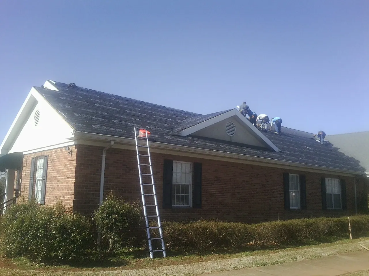 Crew tearing shingles off a brick ranch roof with a ladder set — Emergency Roof Repair work by our crew in Ninety Six