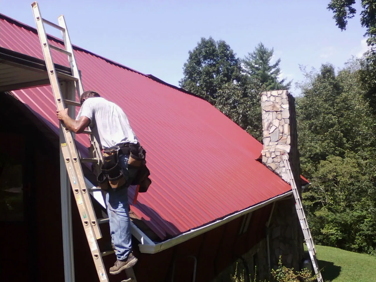 Roofer on a ladder beside a red metal roof and stone chimney — Soffit & Fascia Repair by Roofing Brothers in Ninety Six